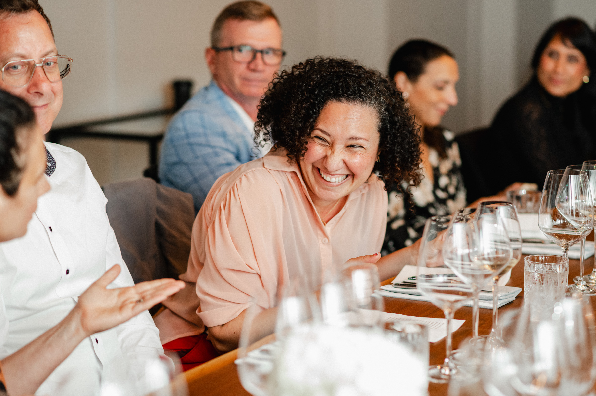 A group of people sits around a table, including a woman with curly hair laughing. Several wine glasses and plates adorn the table, capturing the essence of joyful moments typically seen in event photography. Others are looking in different directions.