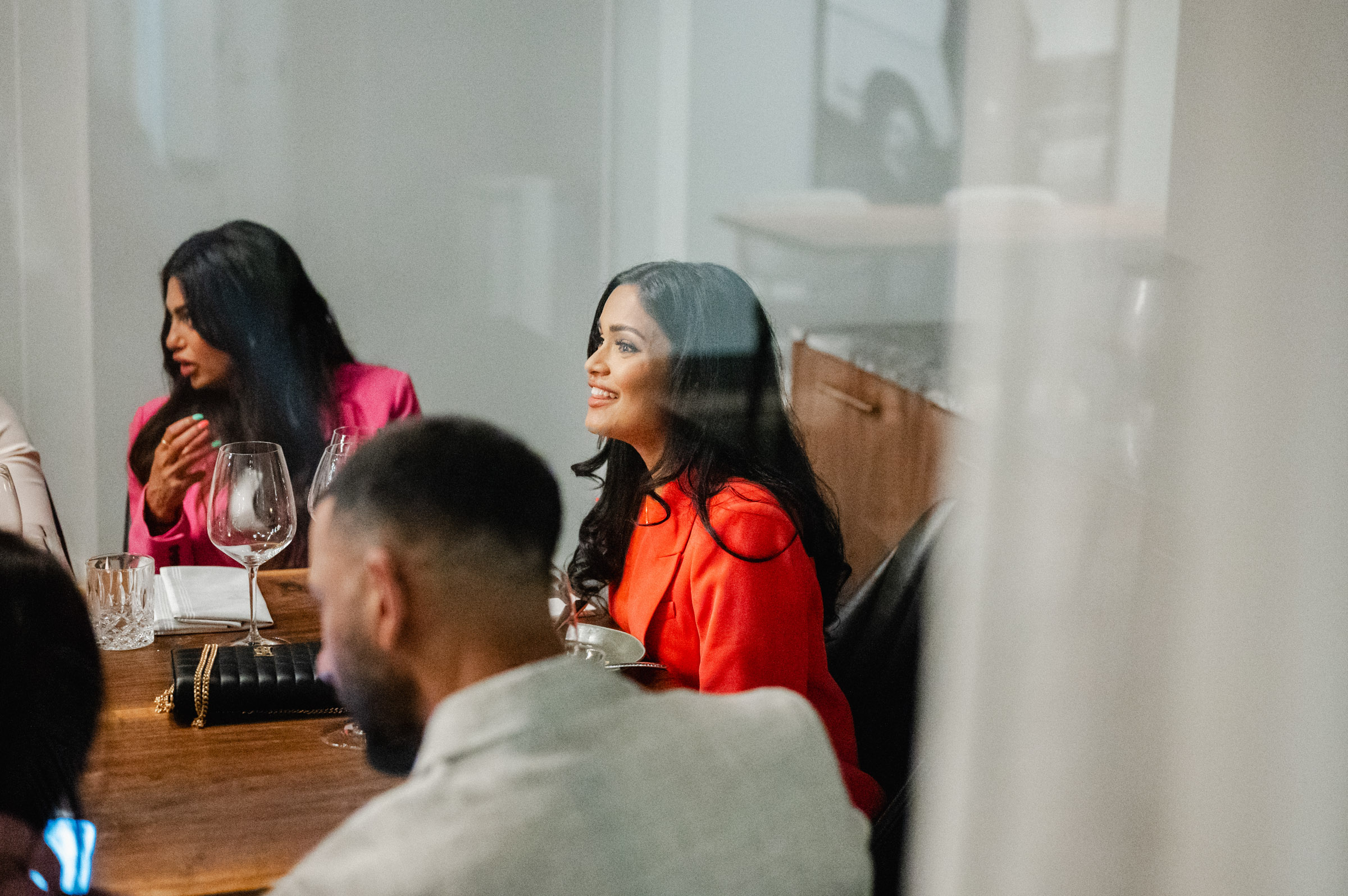 Three people sit around a table, with one woman in a bright red outfit smiling. Wine glasses and plates are visible on the table. The image, captured through a glass window, exemplifies event photography at its finest.