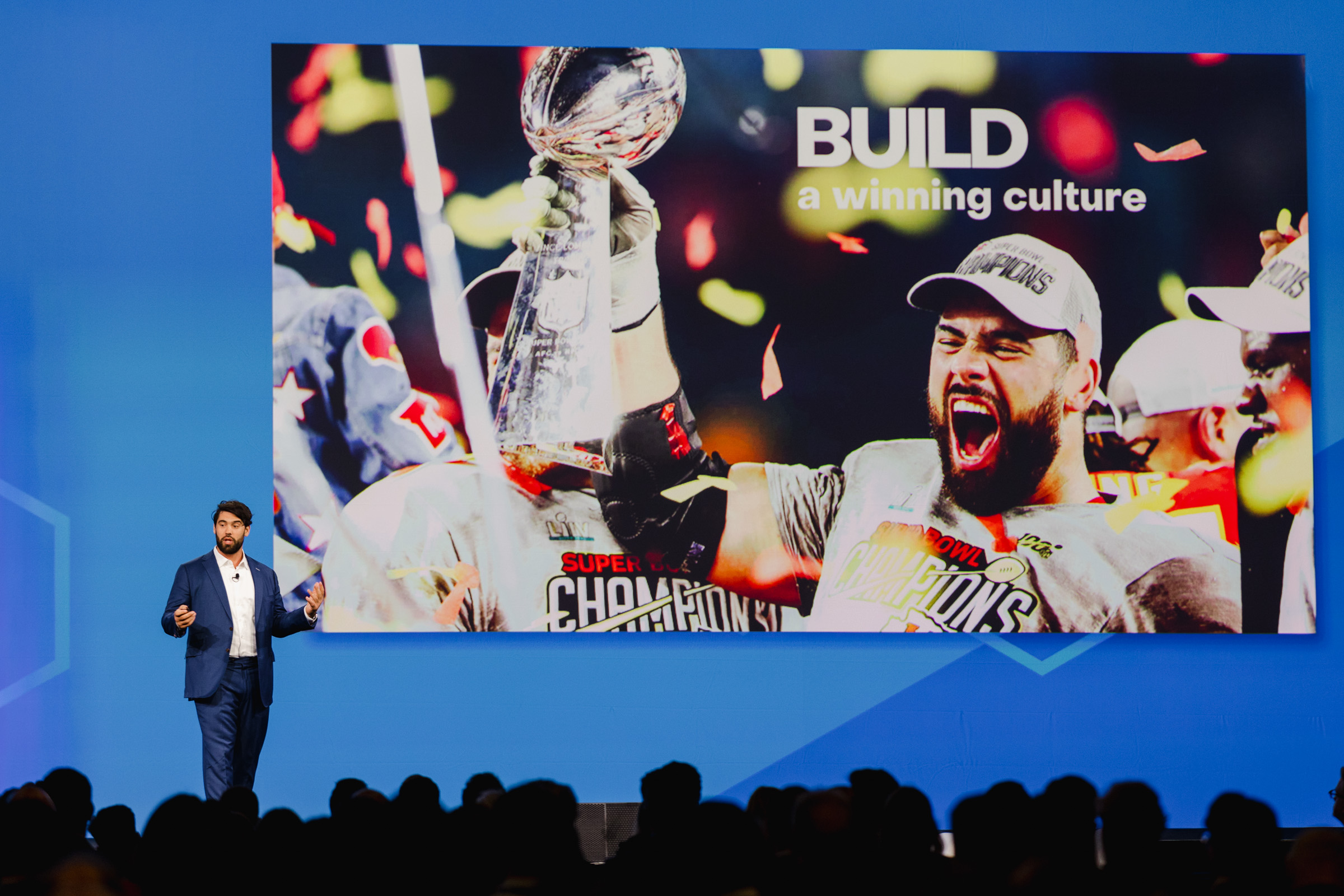 A speaker presents on stage, with event photography capturing a large screen behind them showing a football player celebrating, holding a trophy. The screen reads "BUILD a winning culture." Audience members are visible in the foreground.