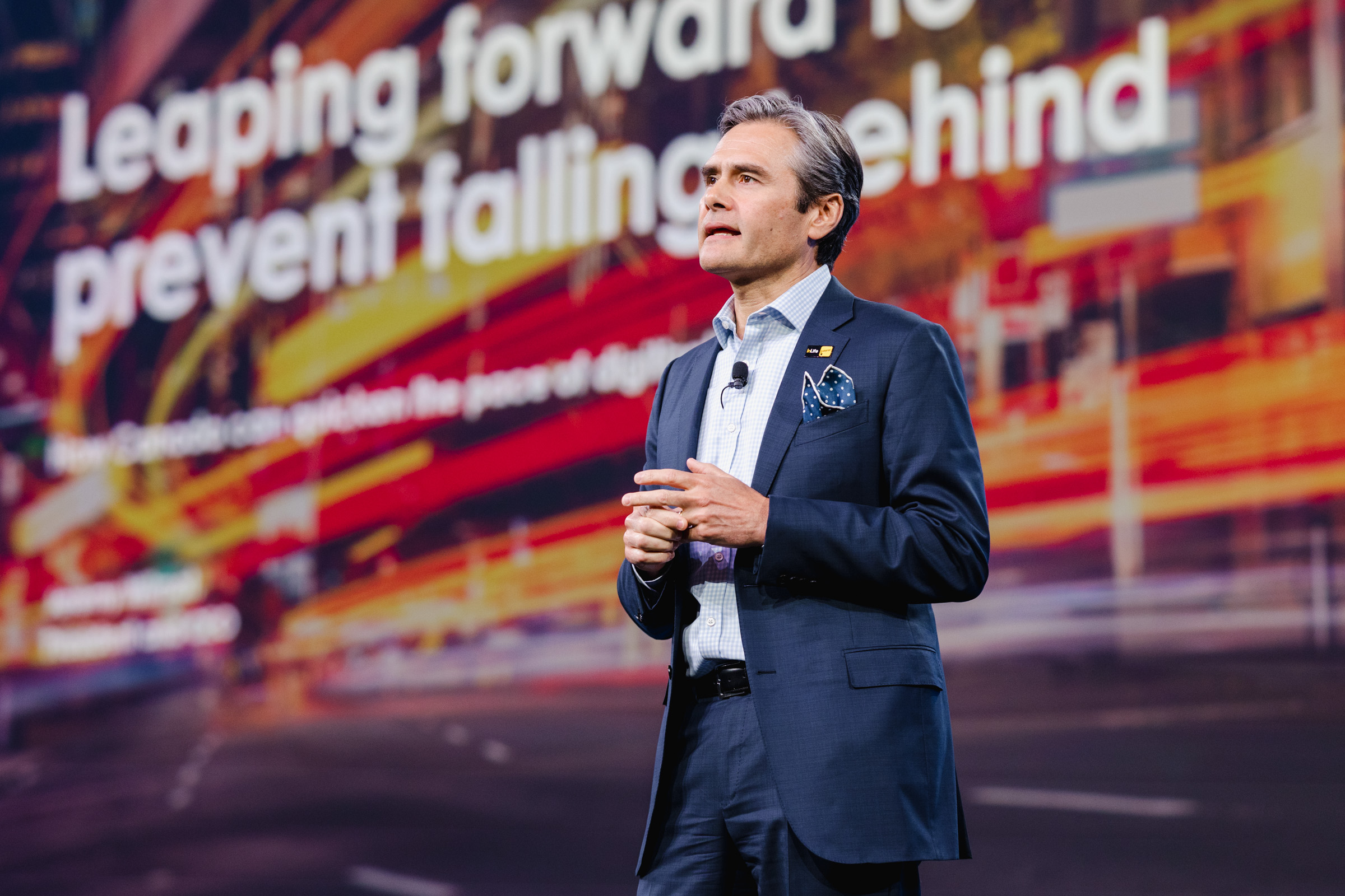 Man in a suit speaking on stage with a blurred background displaying the text, "Leaping forward to prevent falling behind." Captured through expert event photography techniques.