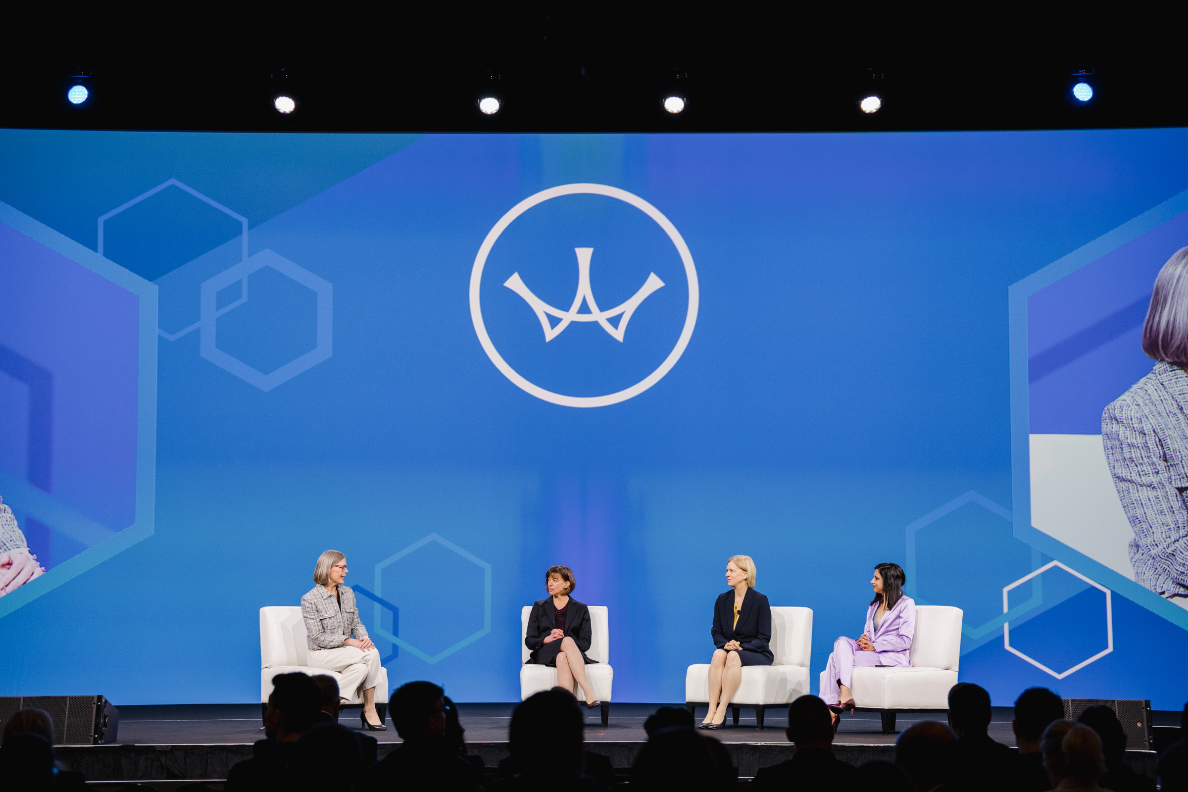 Four individuals are seated on stage, participating in a panel discussion. The background features a large logo and geometric shapes on a blue screen. Capturing the moment with event photography, an audience is visible in the foreground.