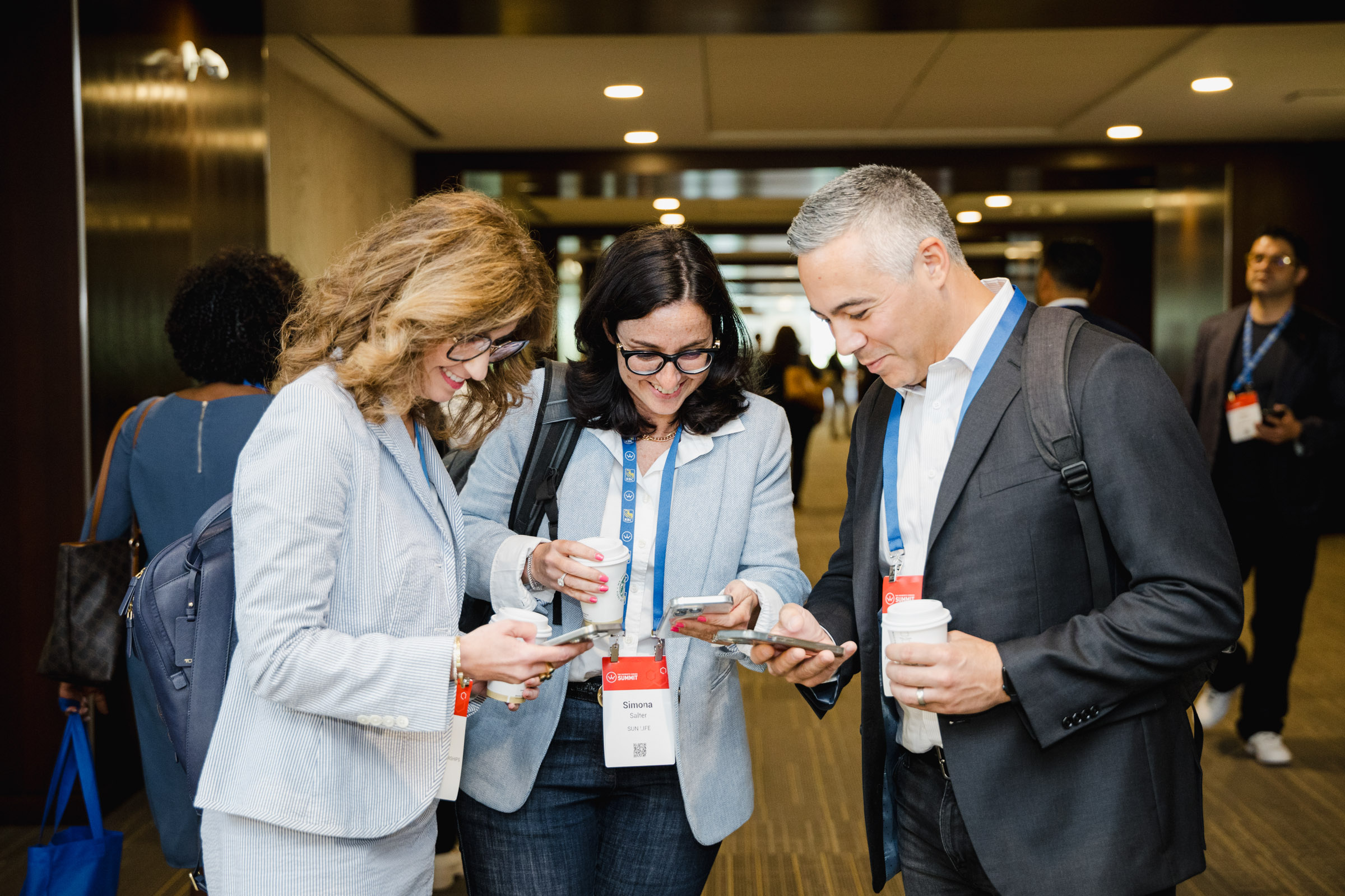 Three individuals in business attire look at their phones while standing together in a hallway. They are wearing name badges and holding coffee cups, capturing a candid moment perfect for event photography.