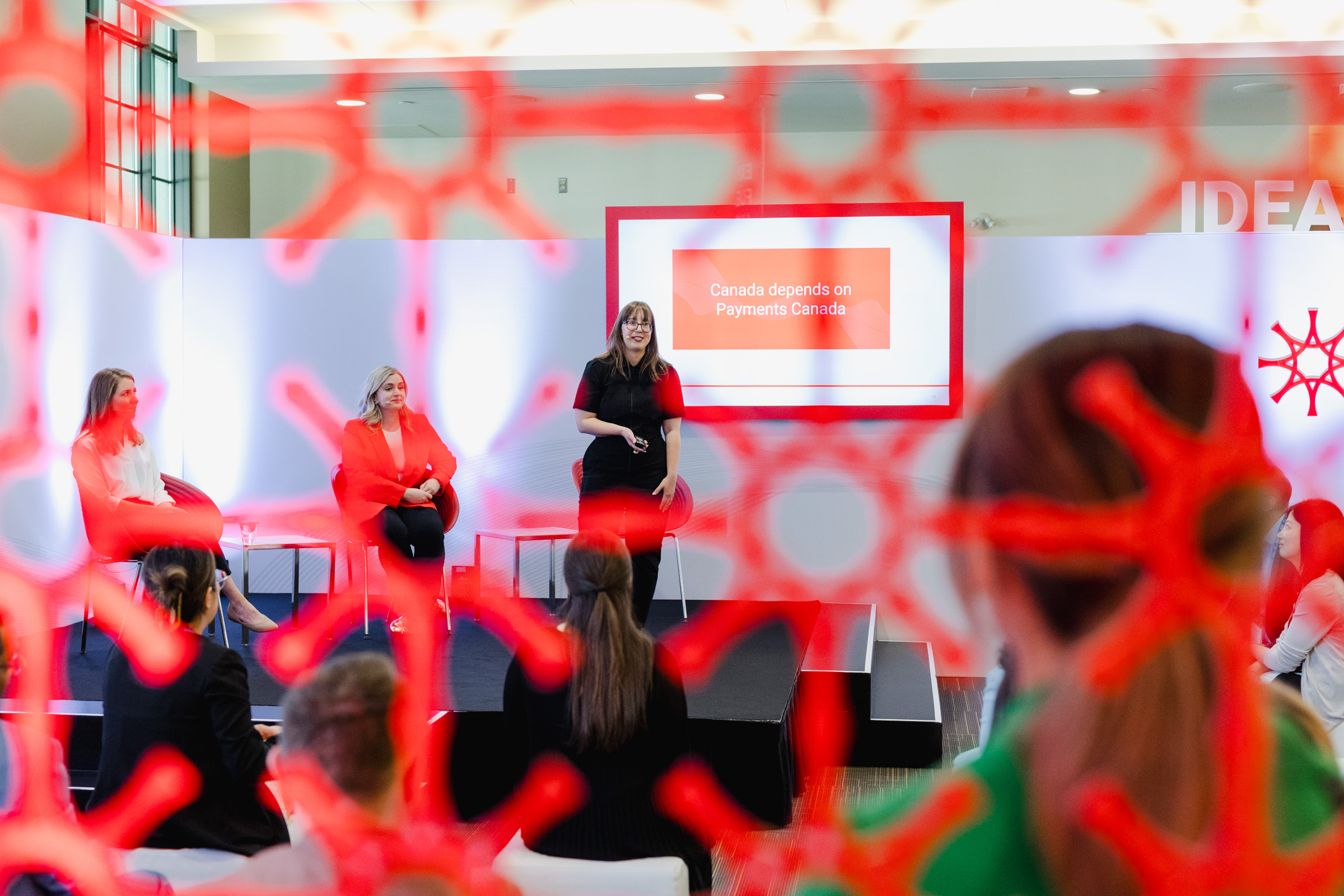 A panel of four people is speaking on a stage at a Payments Canada event, captured with skilled event photography. A screen behind them displays the text, "Canada depends on Payments Canada." The foreground is partially obscured by red netting.