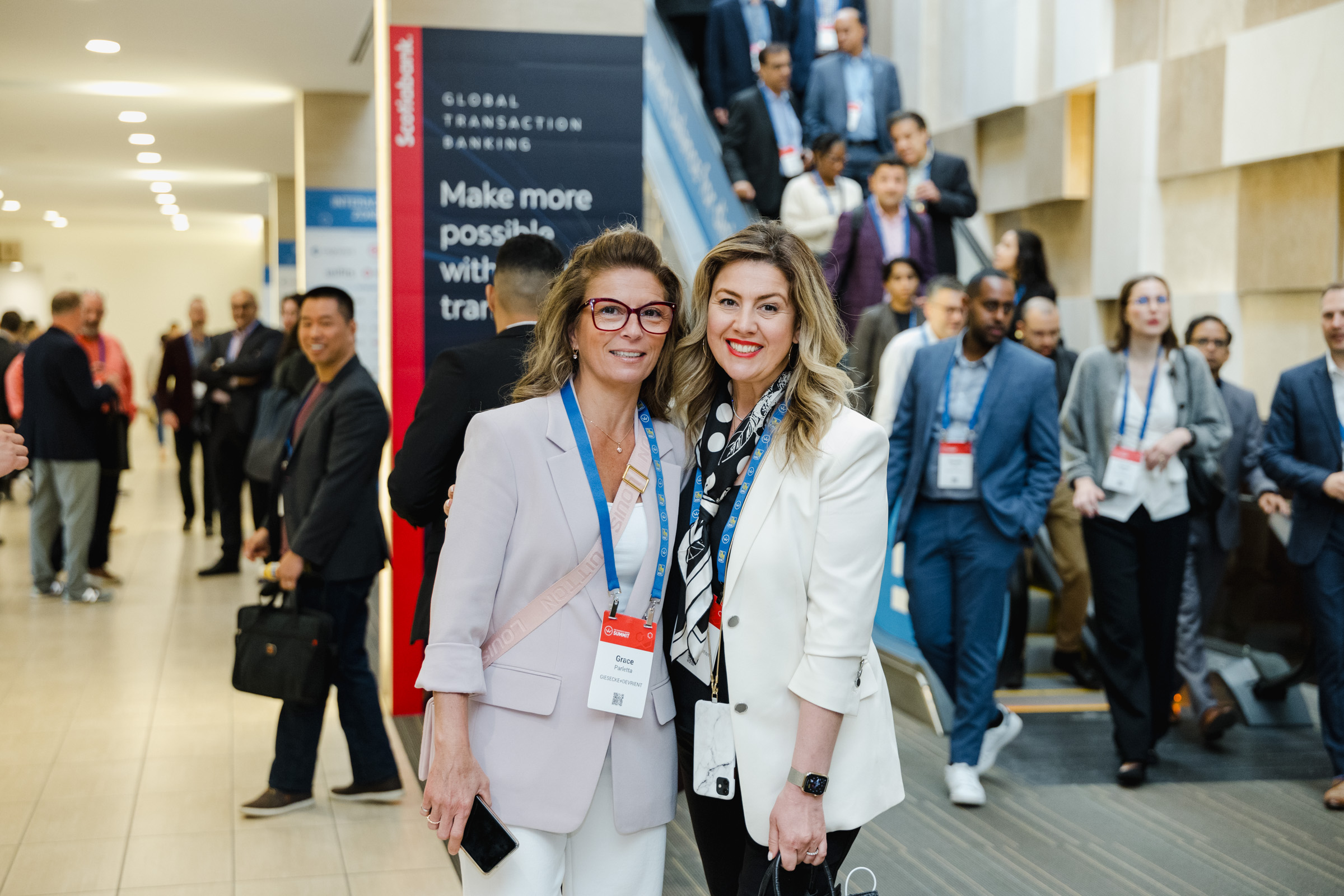 Two women in business attire pose for a photo at a professional event, captured by expert event photography, with a group of people and a sign for 'Global Transaction Banking' visible in the background.