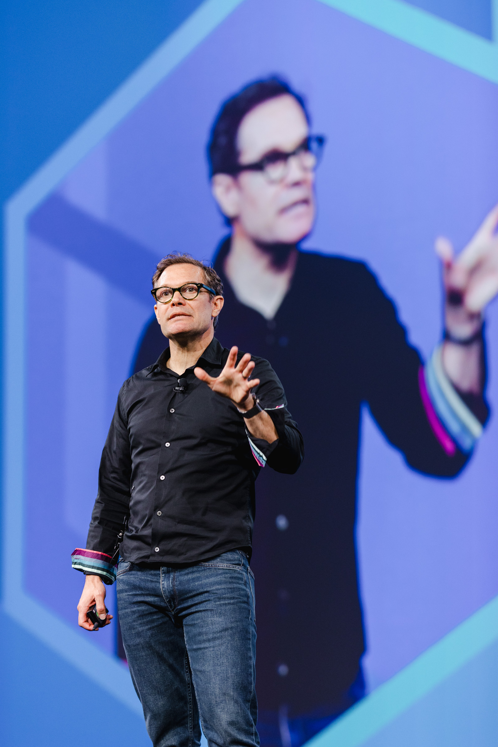 Man in black shirt and jeans speaking on stage, captured through corporate photography, with a large screen displaying his image.