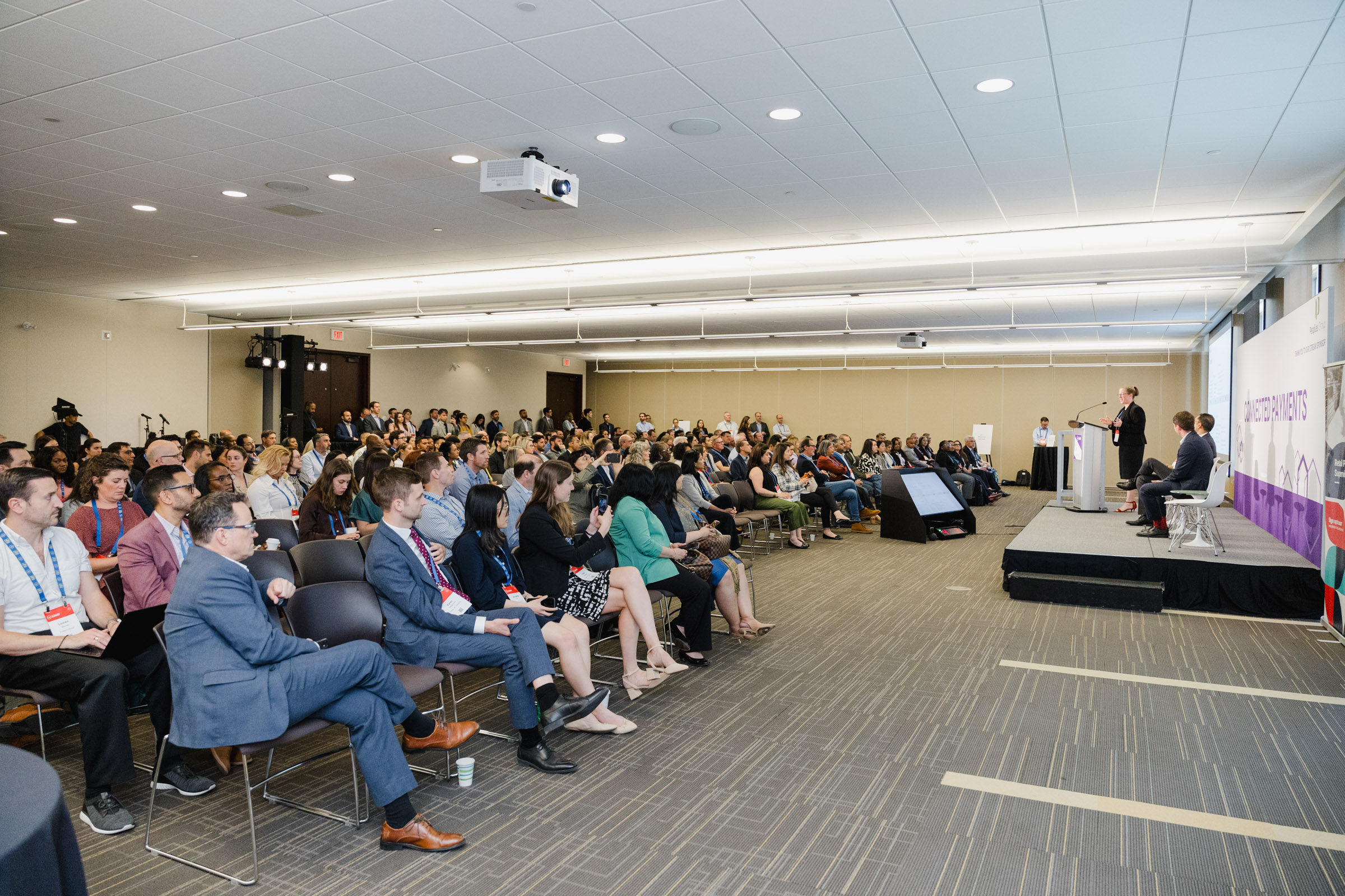A large audience seated in a modern conference room listens to a panel of speakers on stage, all captured through skilled event photography. The room is well lit with a projector on the ceiling and a presentation screen in the background.