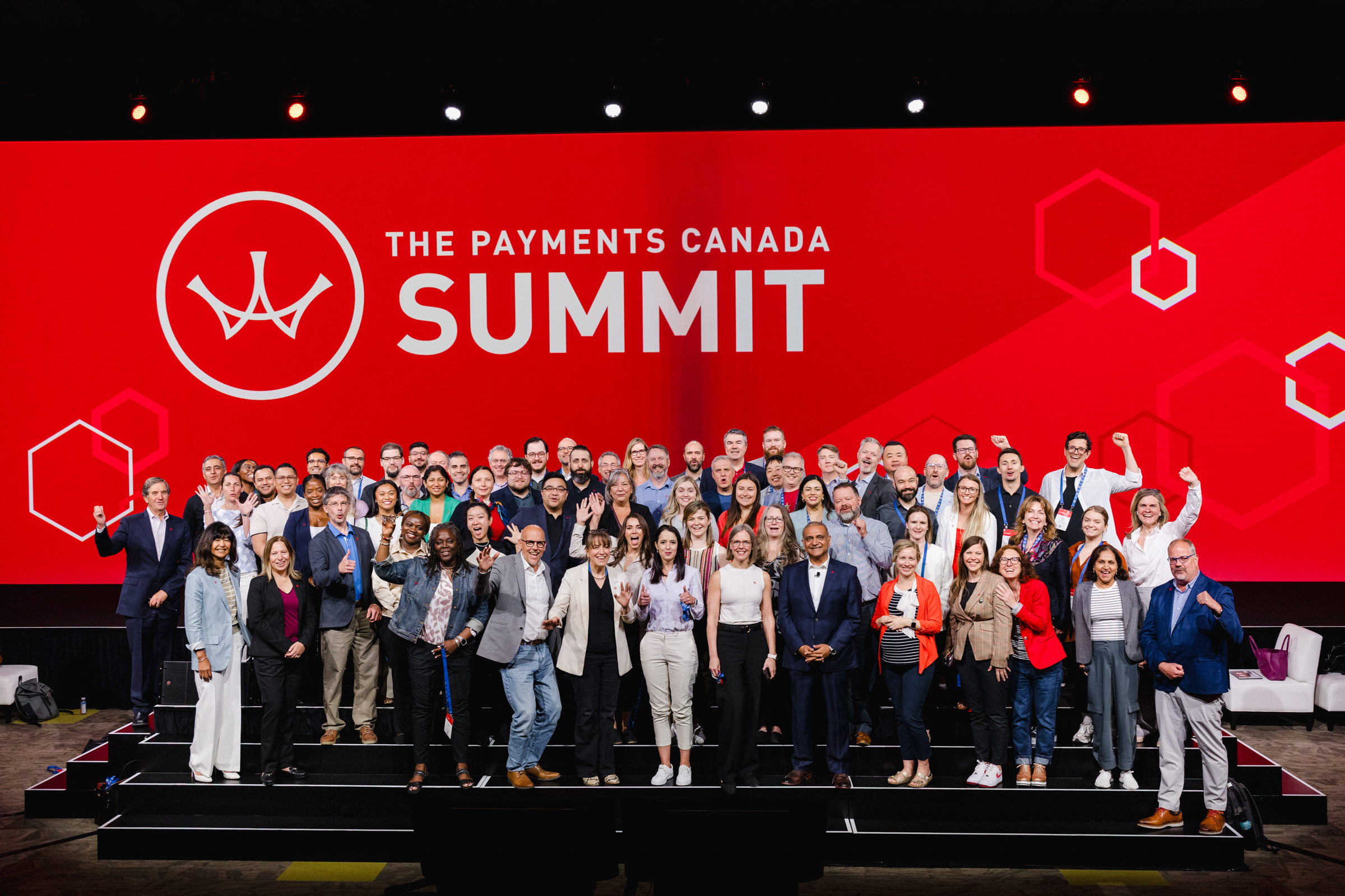 A diverse group of people poses on stage in front of a red backdrop that reads "The Payments Canada Summit," captured perfectly in corporate photography style.