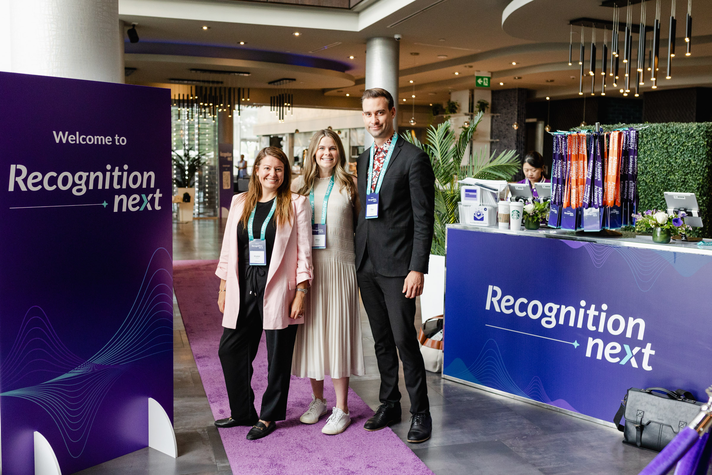 Three people posing in front of a "Recognition Next" conference sign and check-in desk in a modern venue with purple and blue decor, captured perfectly through event photography.