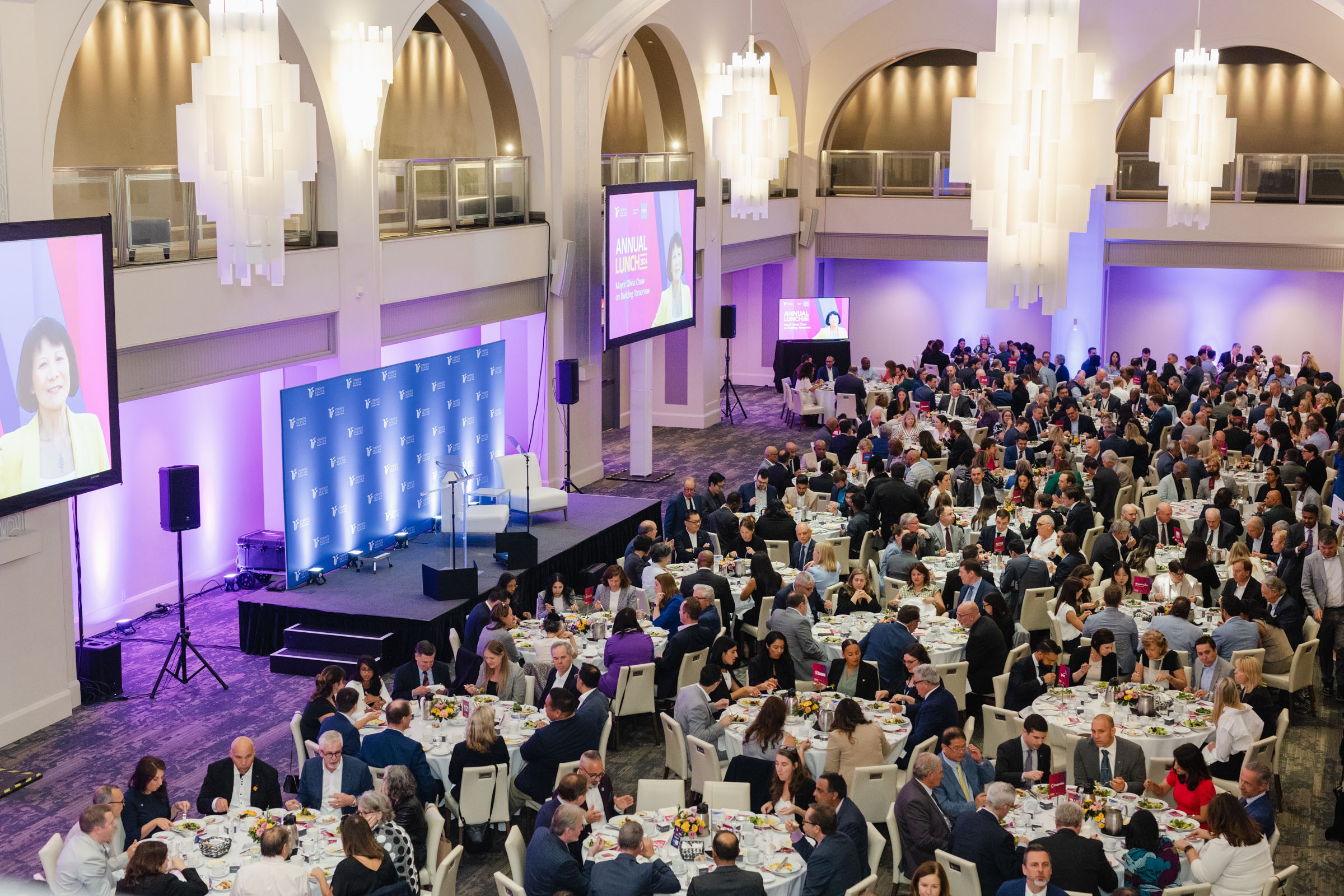 A large group of people attend a formal event in a spacious hall. Tables are set with food, and multiple screens display a speaker, while event photography captures candid moments throughout the evening.