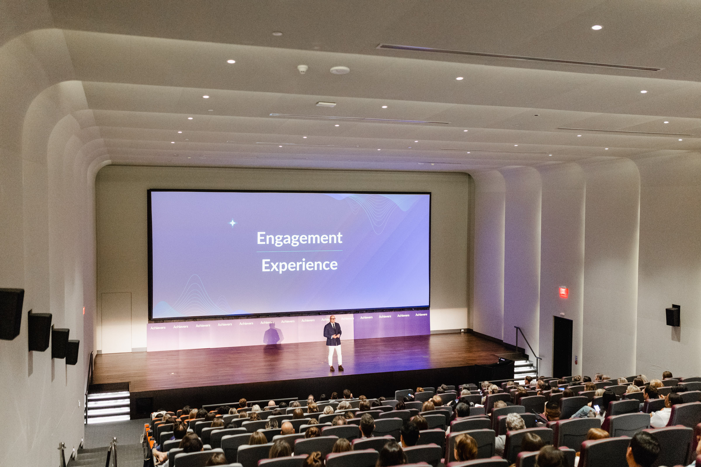 A speaker on stage addresses an audience in a modern lecture hall, with a large screen displaying the words "Engagement" and "Experience." The event photography captures every moment, from the speaker’s passion to the audience's reactions.