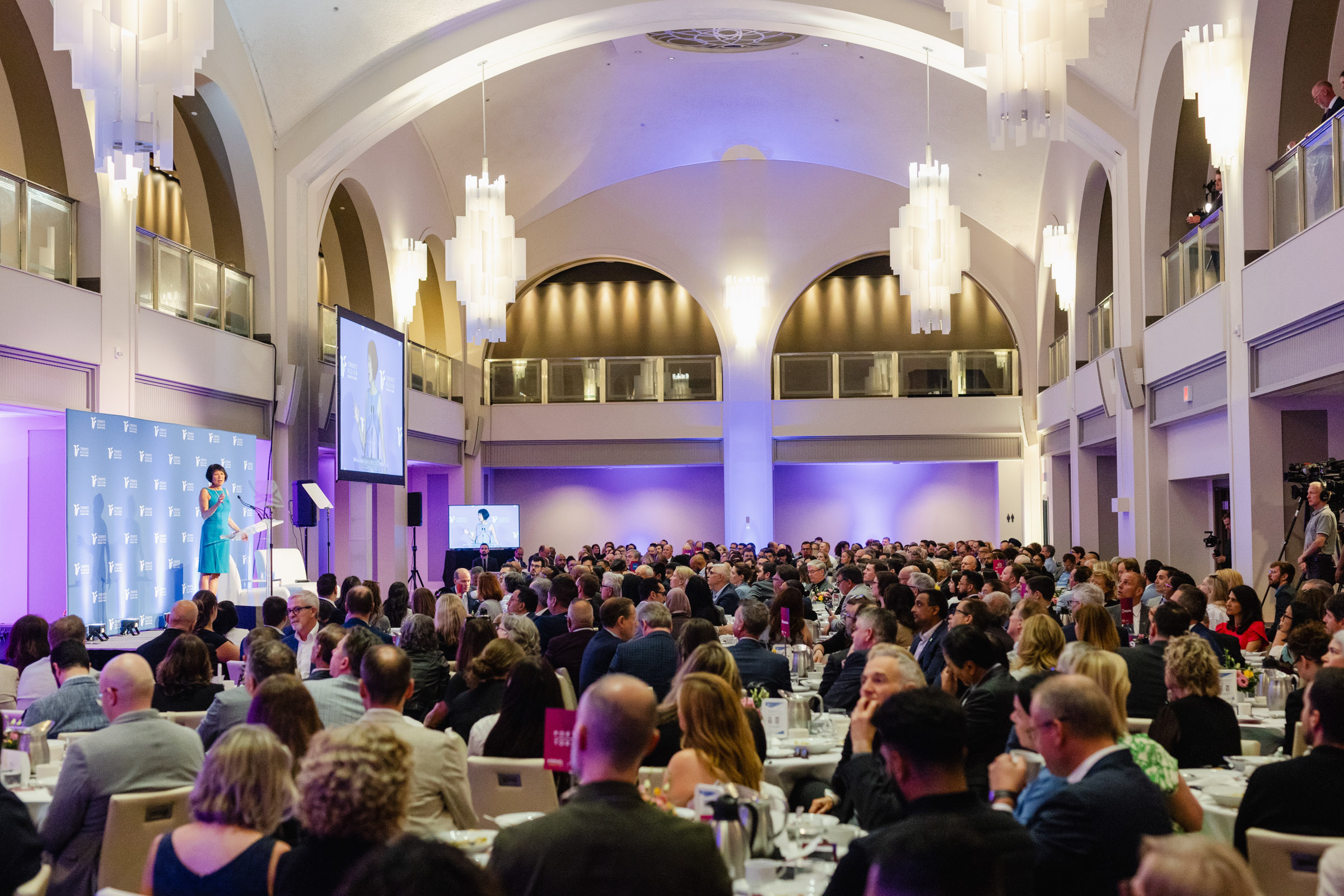 A large audience sits at tables in an elegant hall, attentively watching a speaker on stage beside a presentation screen. The room features high, arched ceilings and modern chandeliers, capturing the perfect setting for event photography.