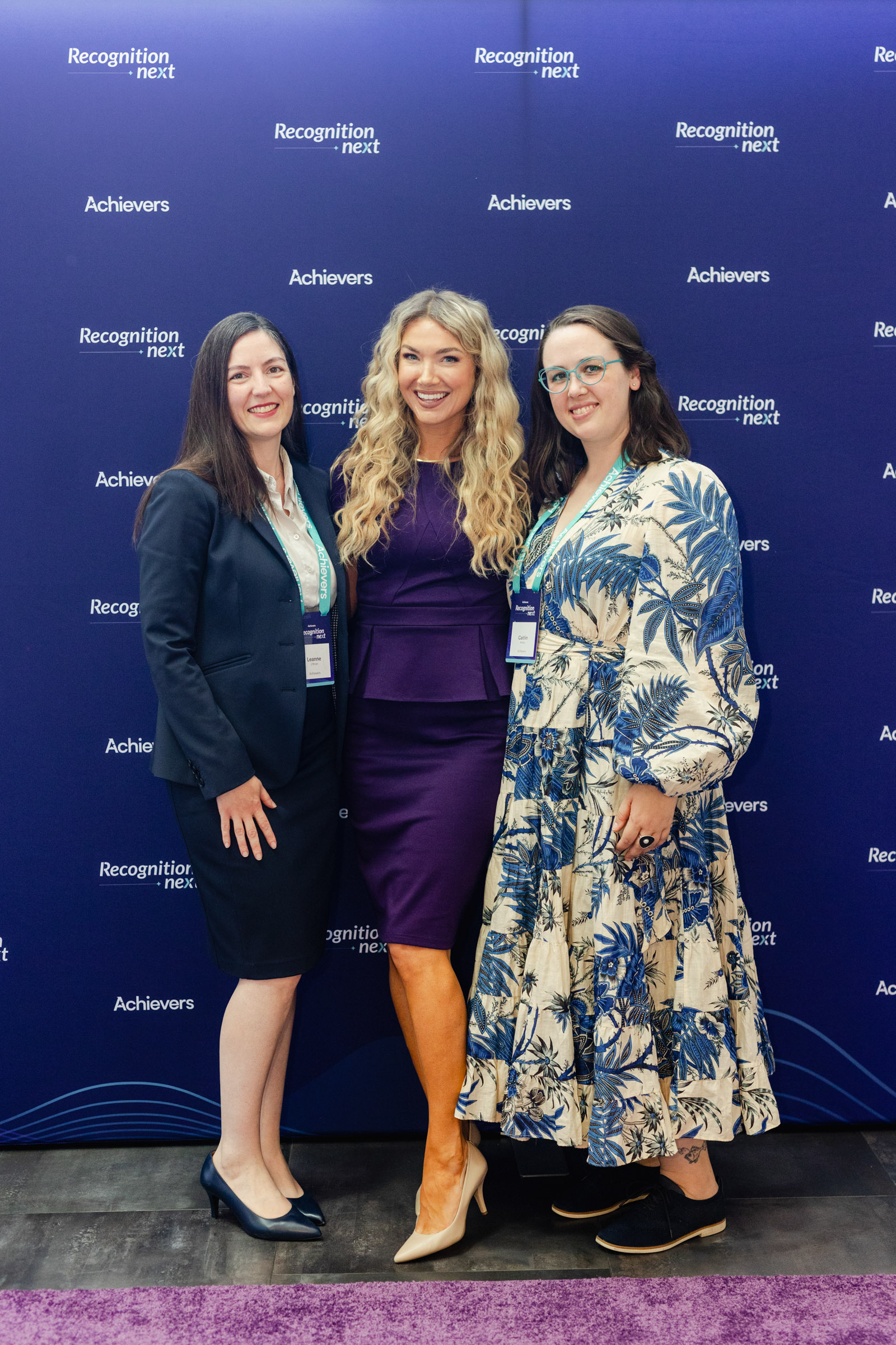 Three women stand in front of a blue "Recognition Next" event backdrop. Two are dressed in business attire, and the third wears a floral dress. All are smiling at the camera, capturing a moment of event photography at its finest.