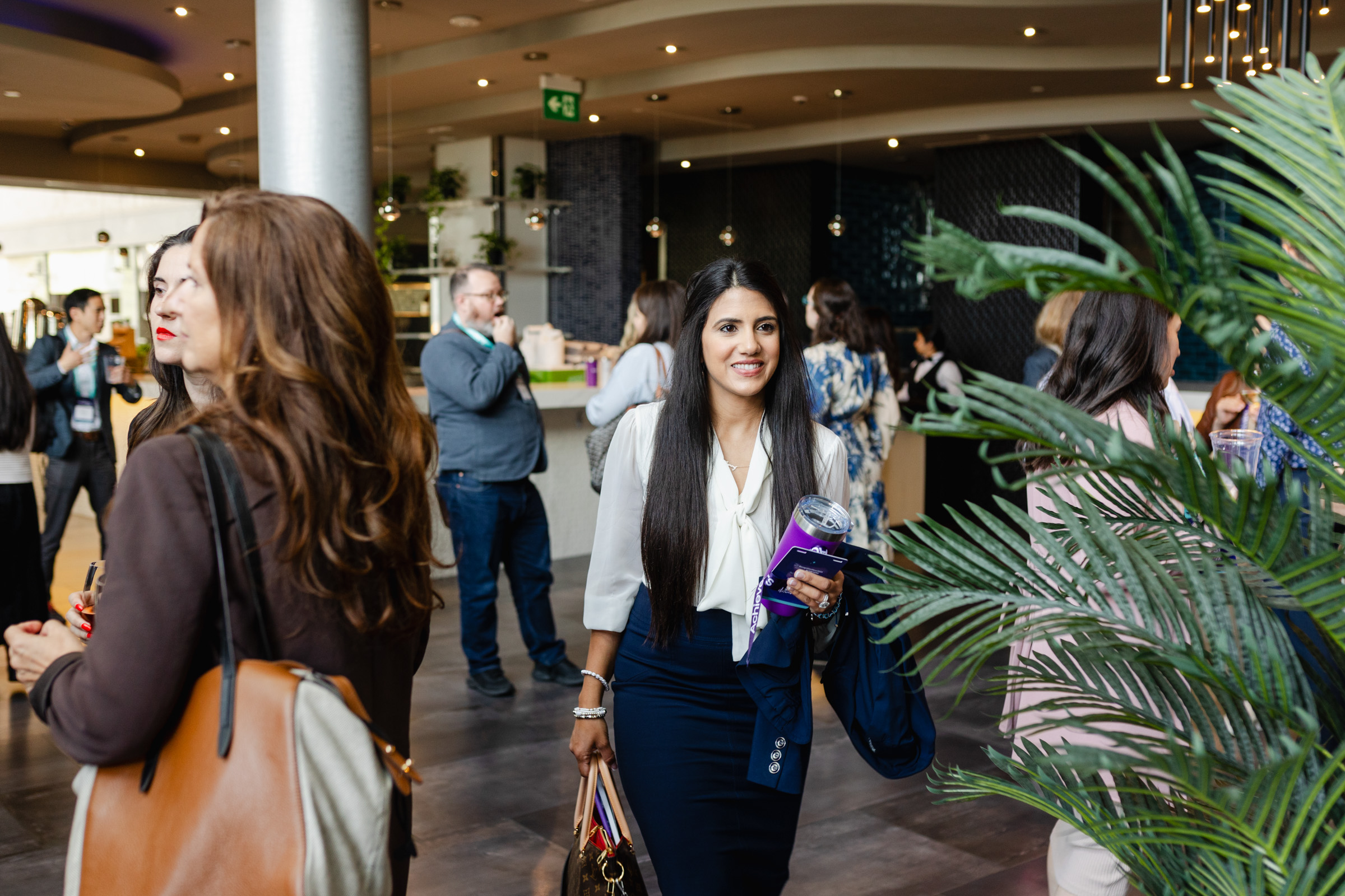 People are attending an indoor networking event. Some individuals are engaged in conversation while others are walking around the space. Event photography captures the scene, with plants and modern decor in the background adding to the atmosphere.