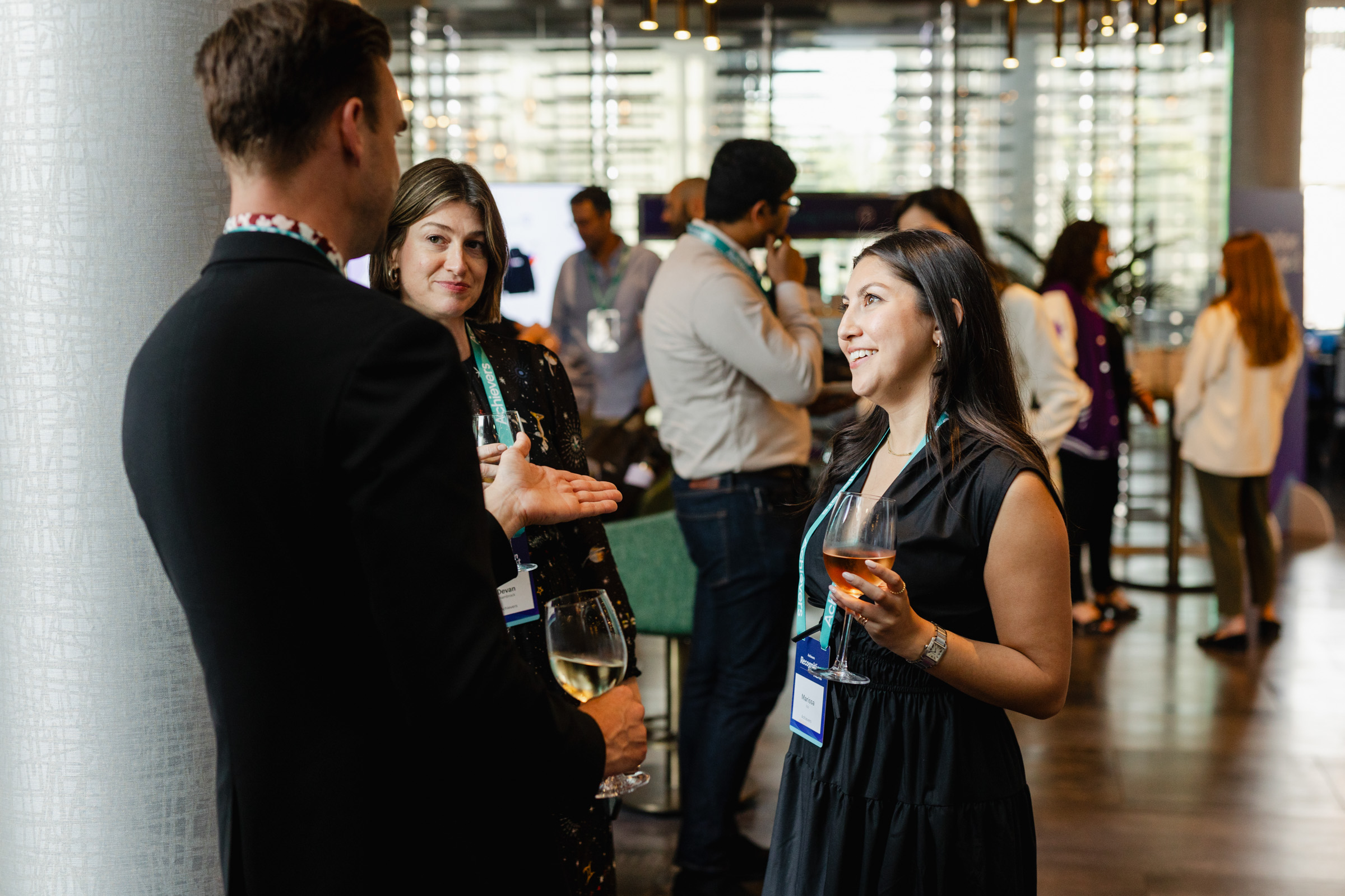 People engaged in conversation at a social event; some hold drinks while wearing name tags. The background features more attendees and a modern interior with large windows, perfectly capturing the essence of event photography.