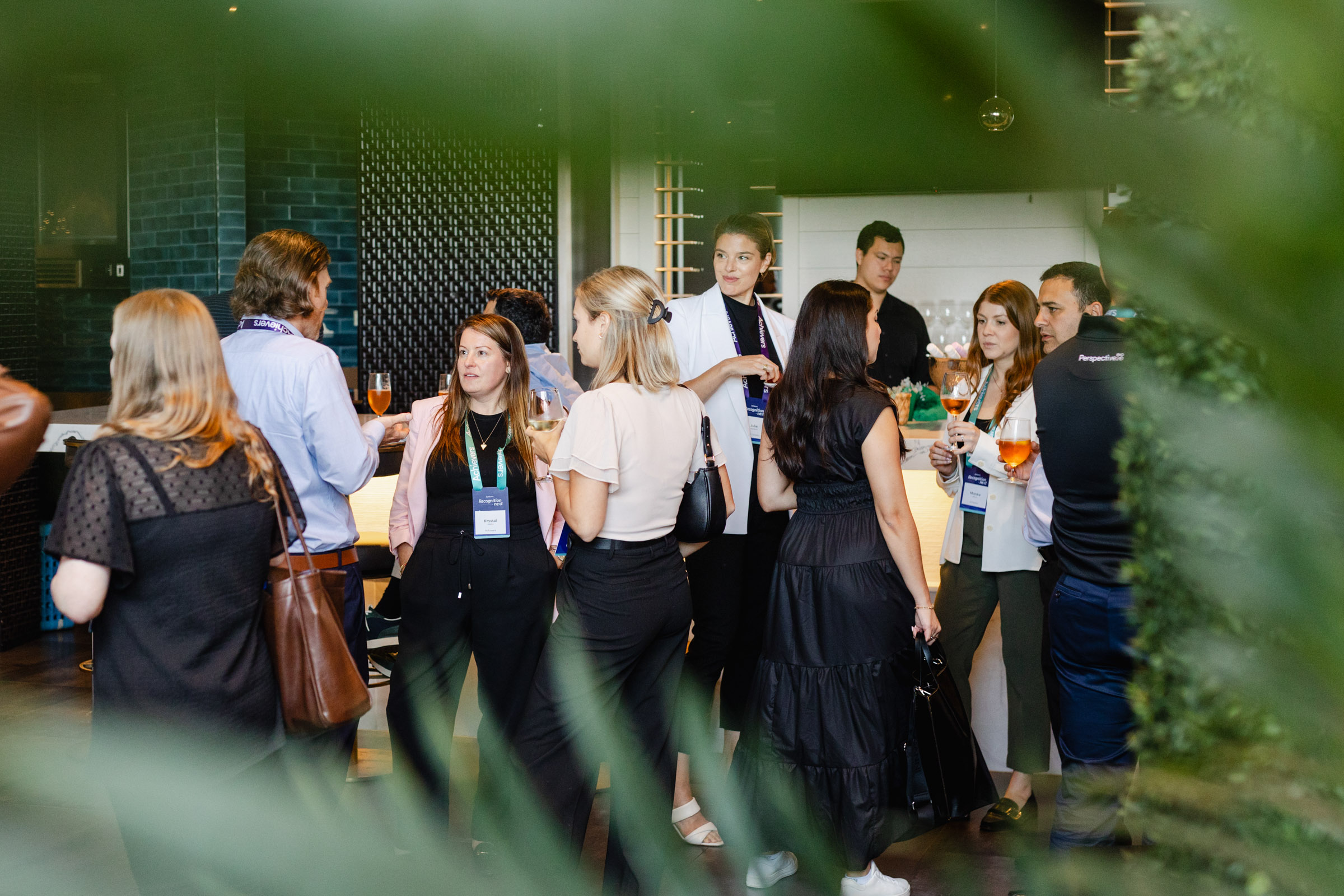 A group of people, dressed in business casual attire, are gathered in a modern indoor space, engaging in conversation while holding drinks. Some individuals, captured through corporate photography, are wearing name badges.