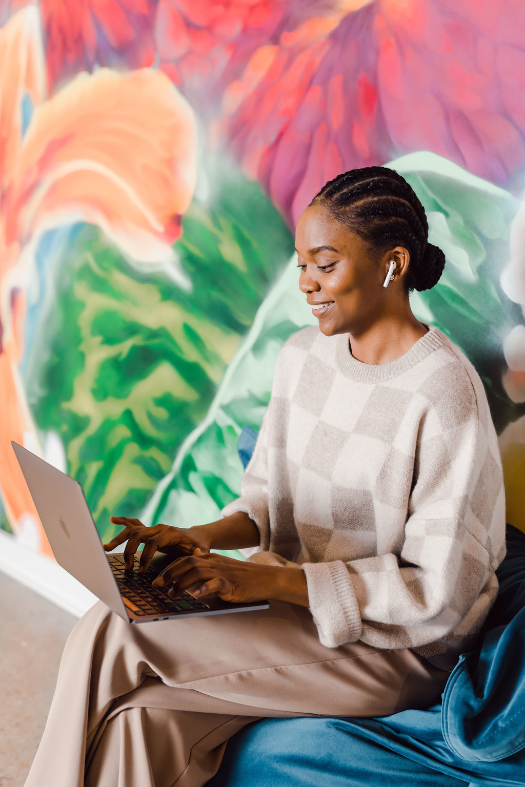A person with braids wearing a checkered sweater and earphones sits on a blue chair, typing on a laptop against a colorful, abstract background, showcasing the creative side of corporate photography.