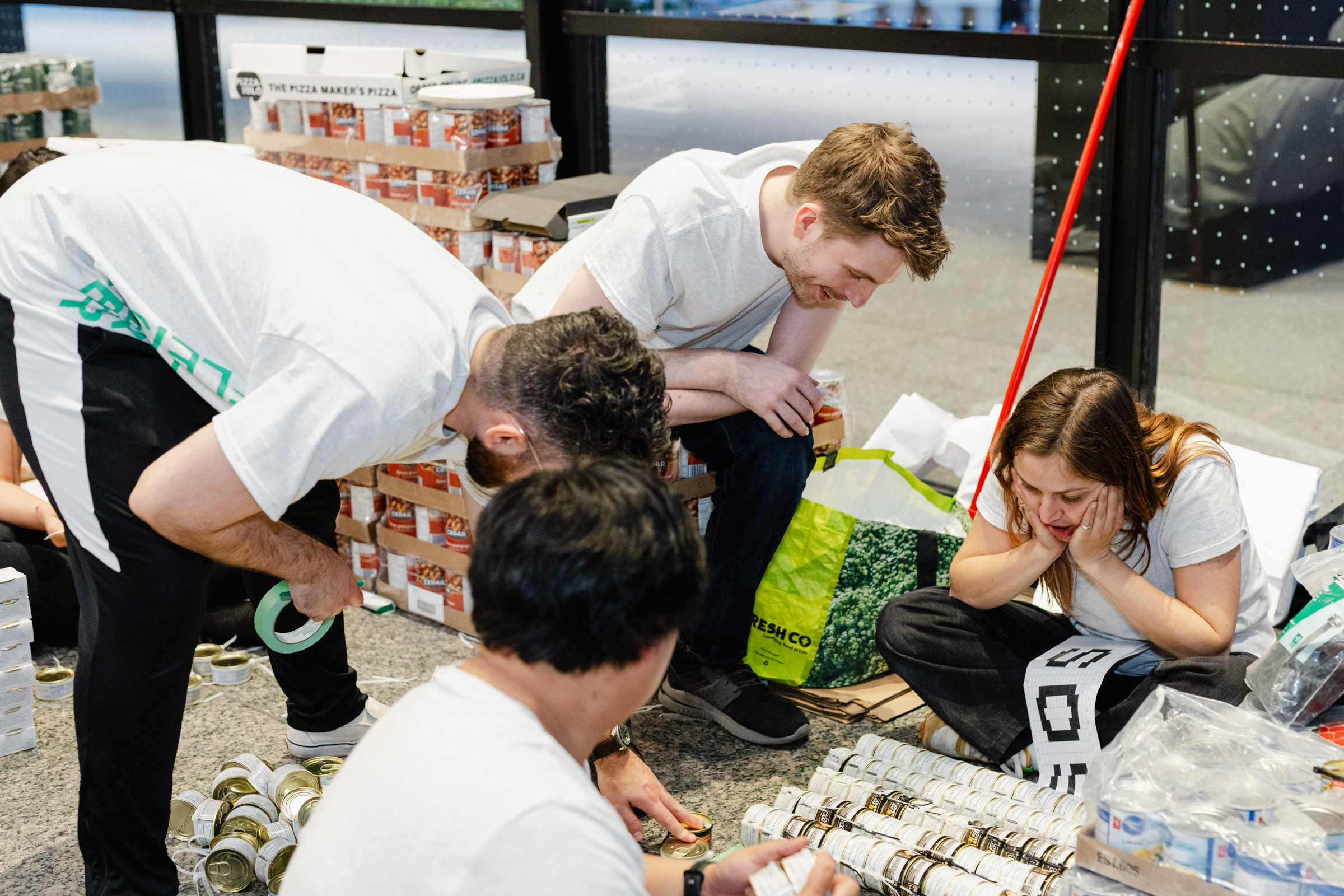 A group of four people sorts and organizes canned food items and boxes in a room. Some are kneeling and some are crouching, engaged in their task, creating a candid scene perfect for event photography.