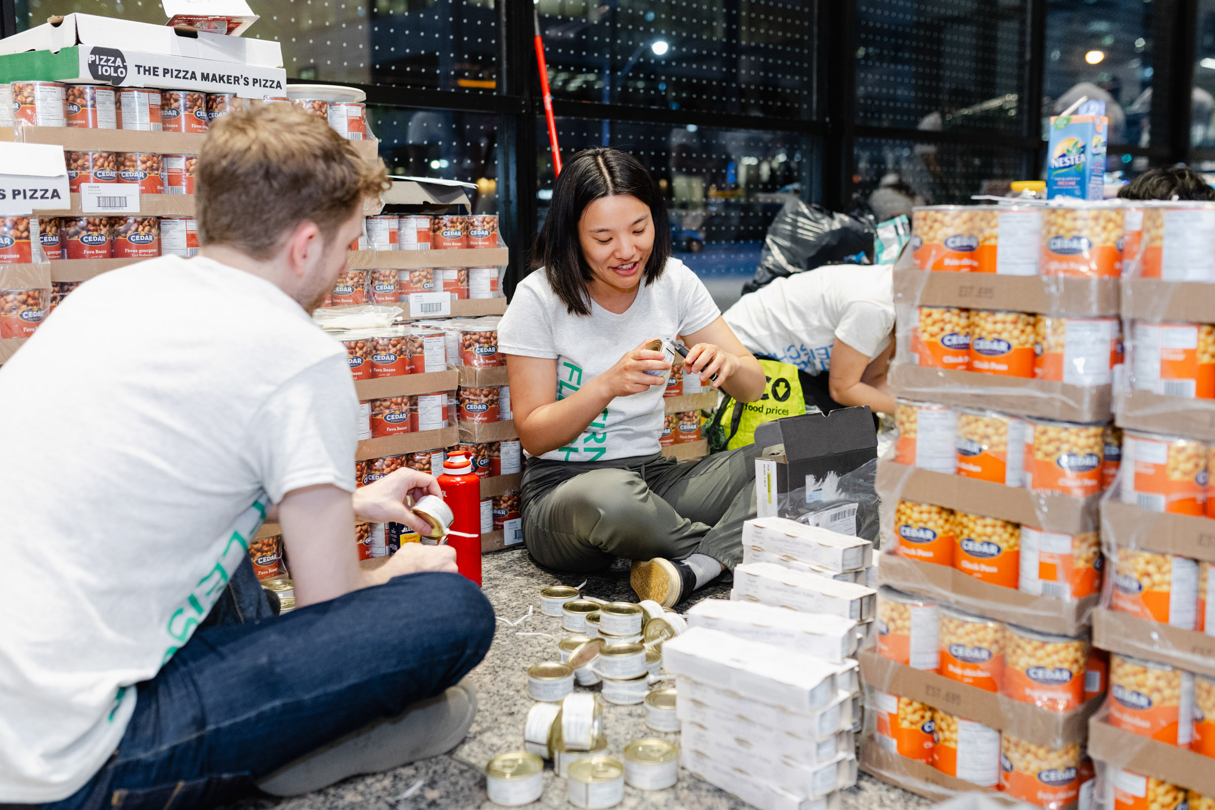 Volunteers seated on the floor organize canned food in a storage area filled with various boxes and stacks of food items, creating a perfect scene for event photography.