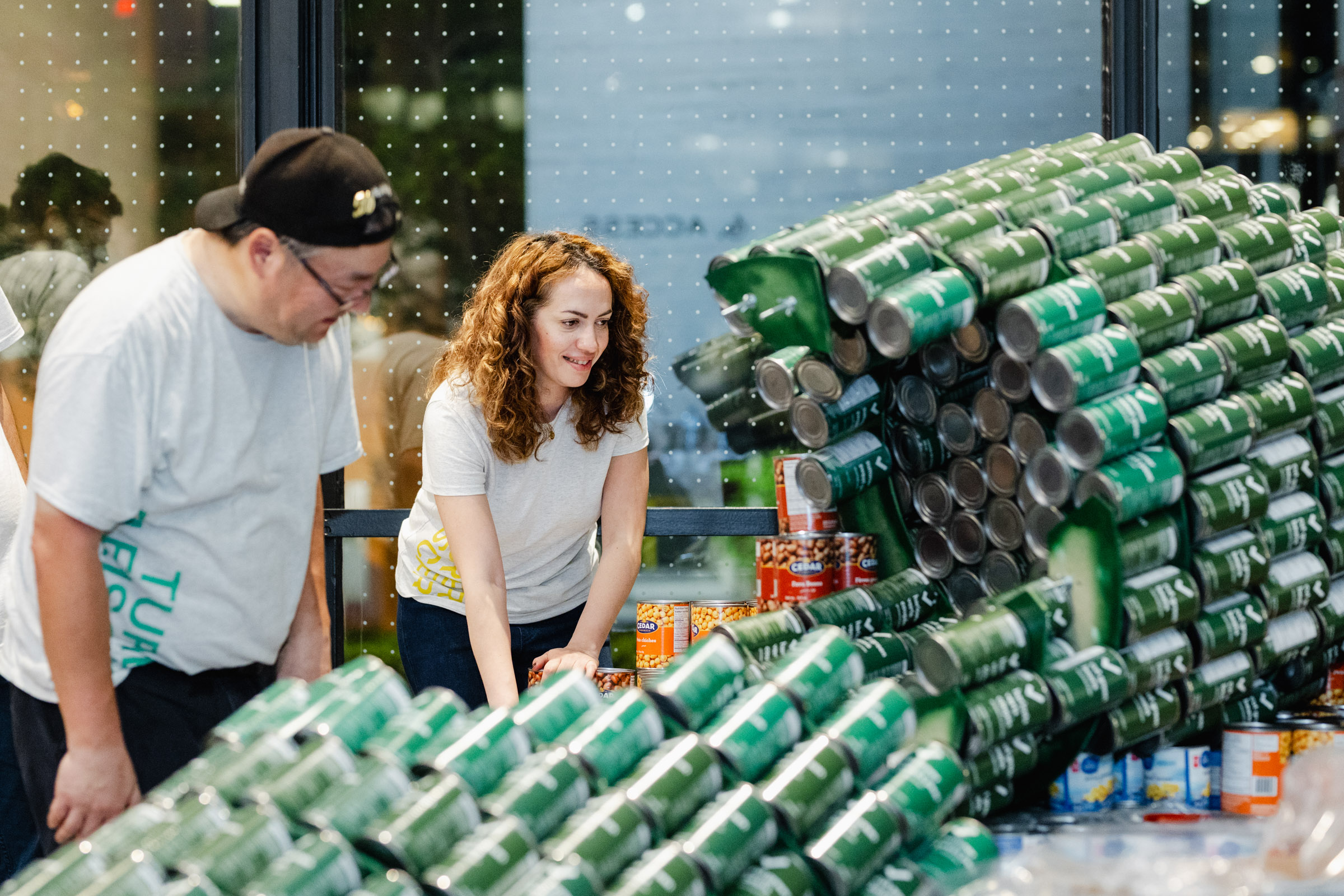 Two individuals are meticulously arranging a large display made of stacked green cans indoors. With the precision of event photography, they seem focused on organizing the structure.