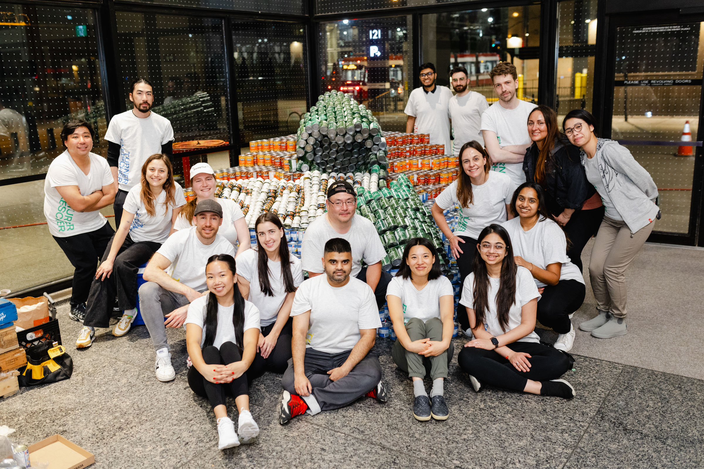 A group of people in white shirts pose together for event photography in front of a large sculpture made of canned goods inside a building.