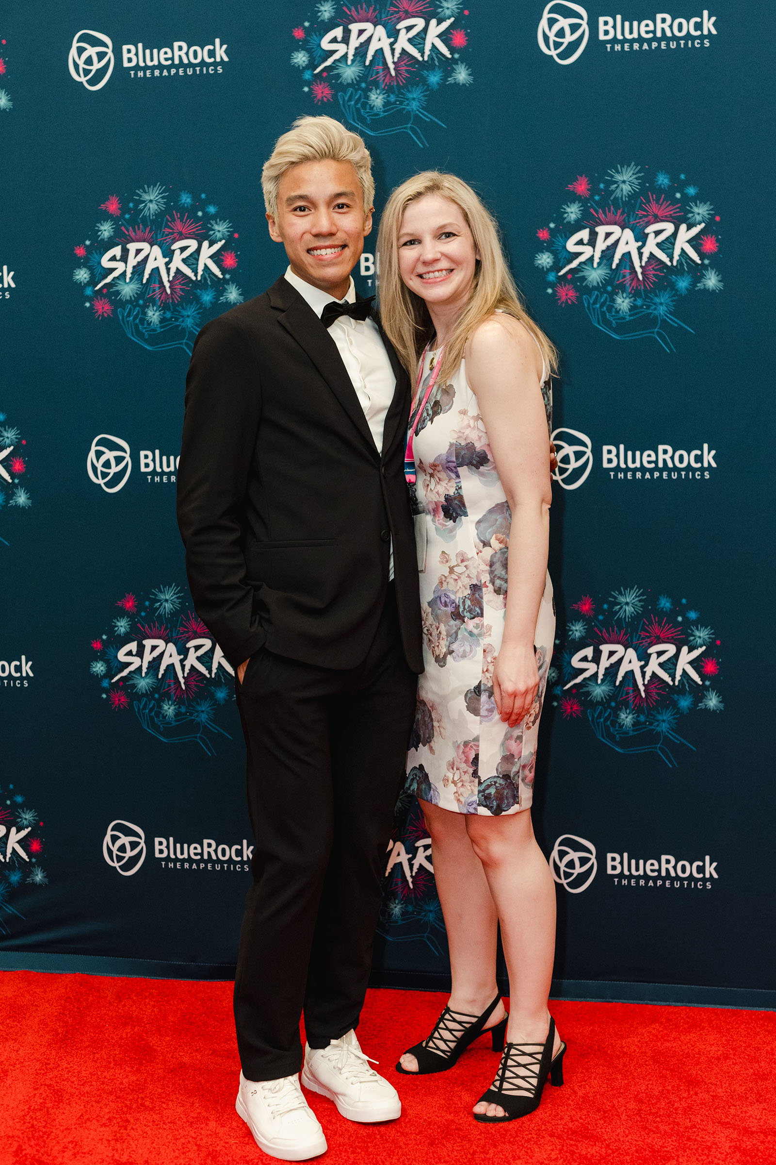 Two people stand on a red carpet in front of a BlueRock Therapeutics "Spark" event backdrop. One person is in a black suit and white sneakers, and the other is in a floral dress and black heels. Event photography captures this stylish moment perfectly.