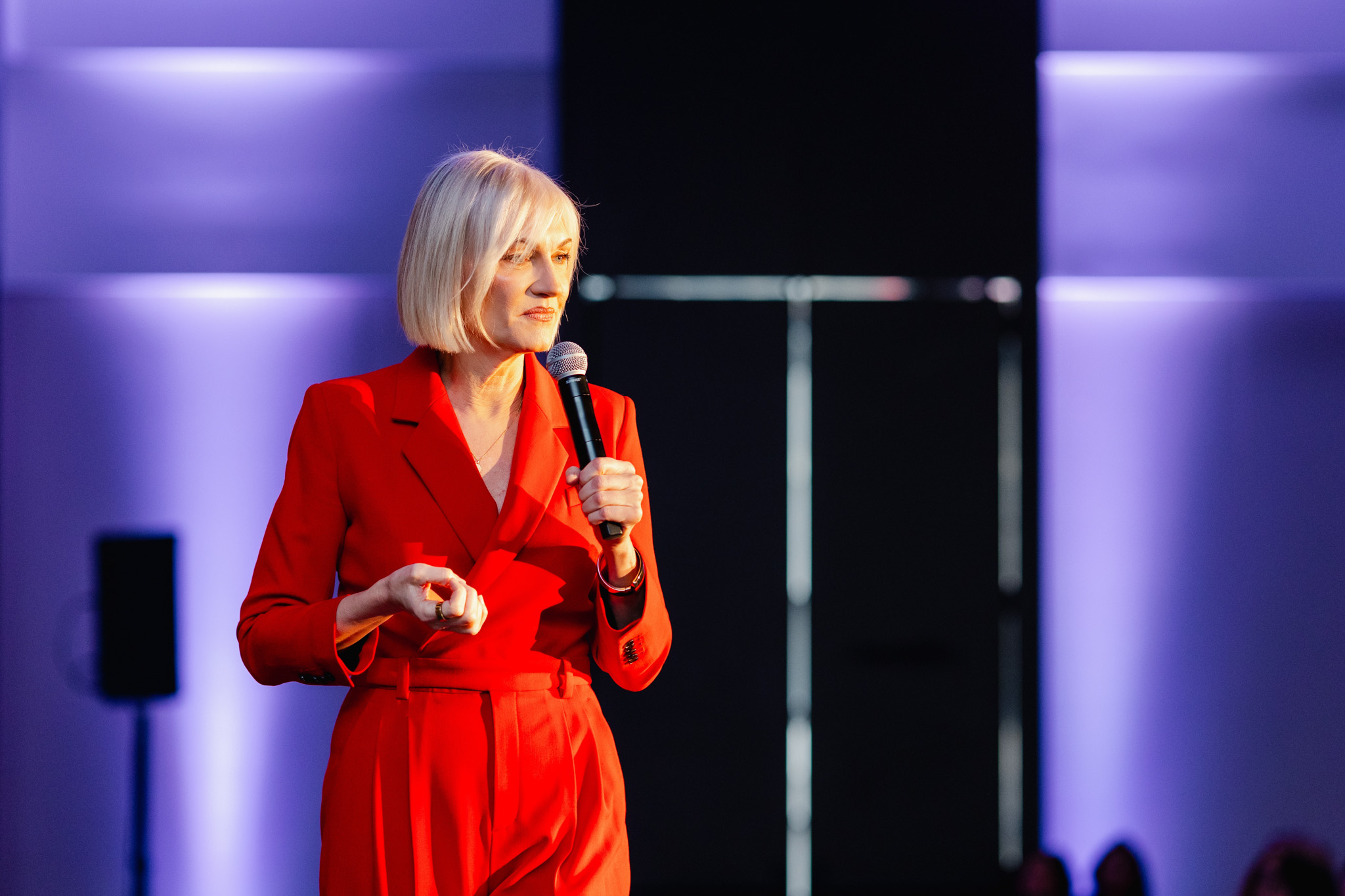 A person with short blonde hair, wearing a red suit, holds a microphone and speaks on stage with purple lighting in the background, capturing the essence of corporate photography.