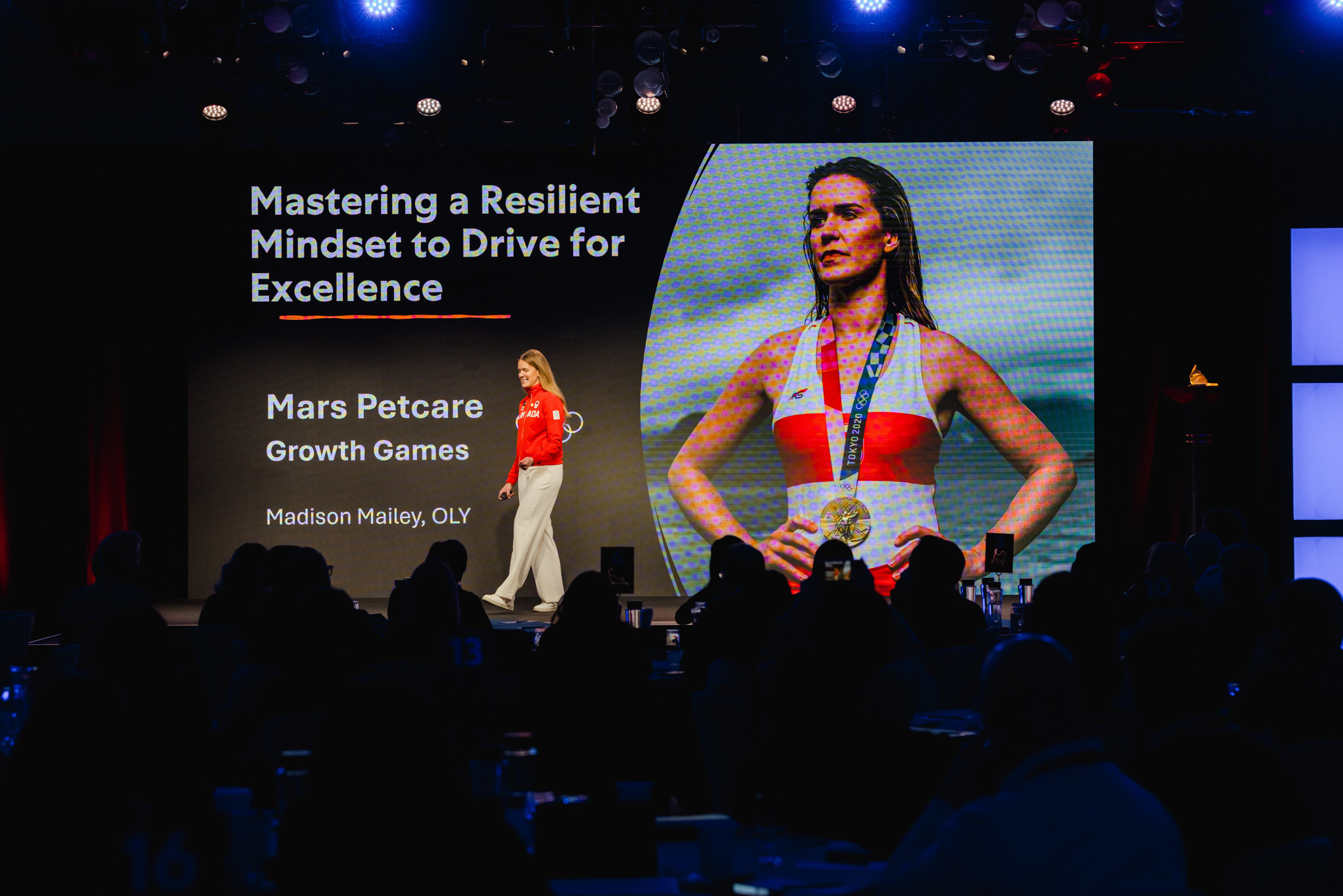 A woman stands on stage presenting in front of an audience with a large screen displaying a female athlete, medals, and the text "Mastering a Resilient Mindset to Drive for Excellence" along with other corporate photography details.