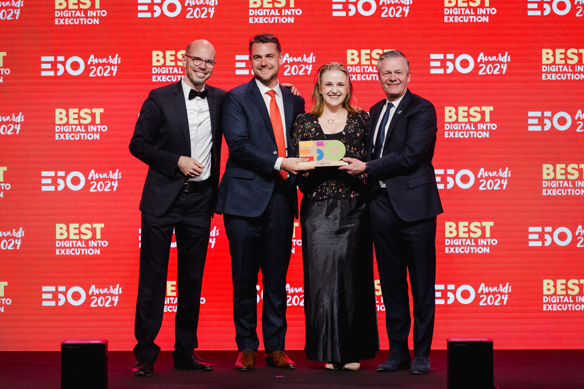 Four people in formal attire stand on stage, smiling and holding an award in front of a red backdrop that reads "Best Digital Into Execution 2024 EO Awards," capturing a moment of corporate photography excellence.