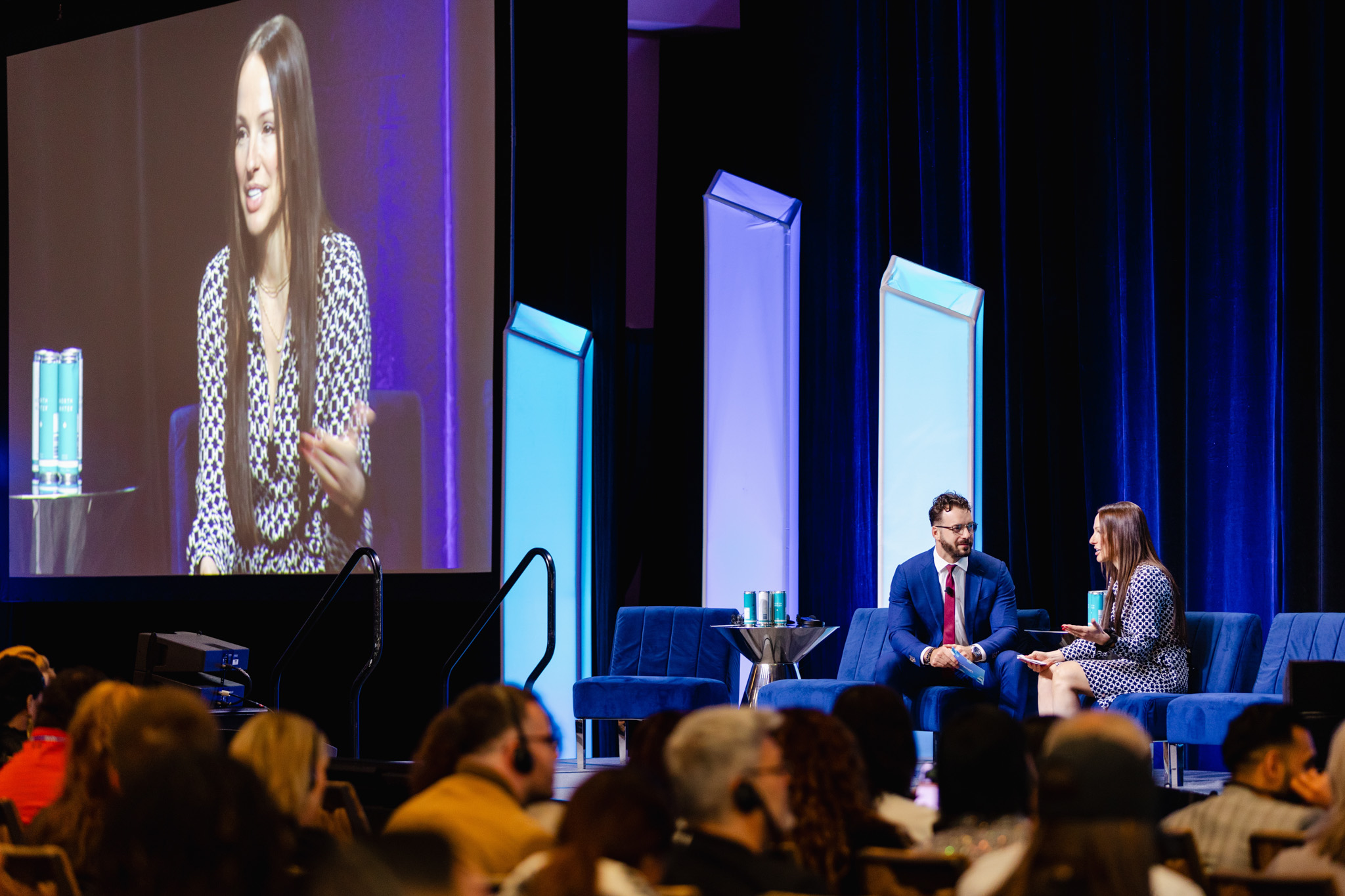Two individuals are seated on a stage engaged in conversation during a conference, perfectly captured through corporate photography. A large screen prominently displays one of the speakers, while an attentive audience is visible in the foreground.