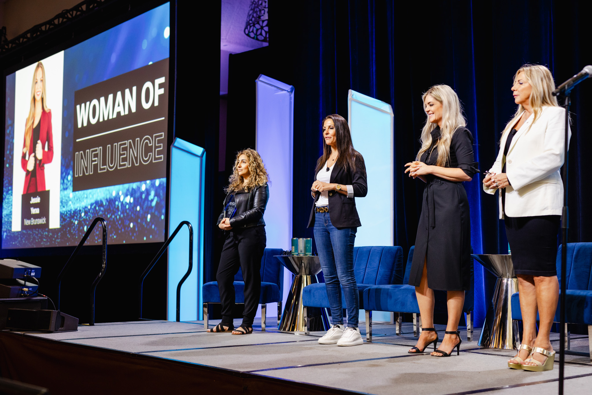 Four women stand on a stage next to a large screen displaying "Woman of Influence." The screen features a portrait of Nicole Arbour, highlighted in an elegant corporate photography style. Chairs and tables are arranged neatly in the background.