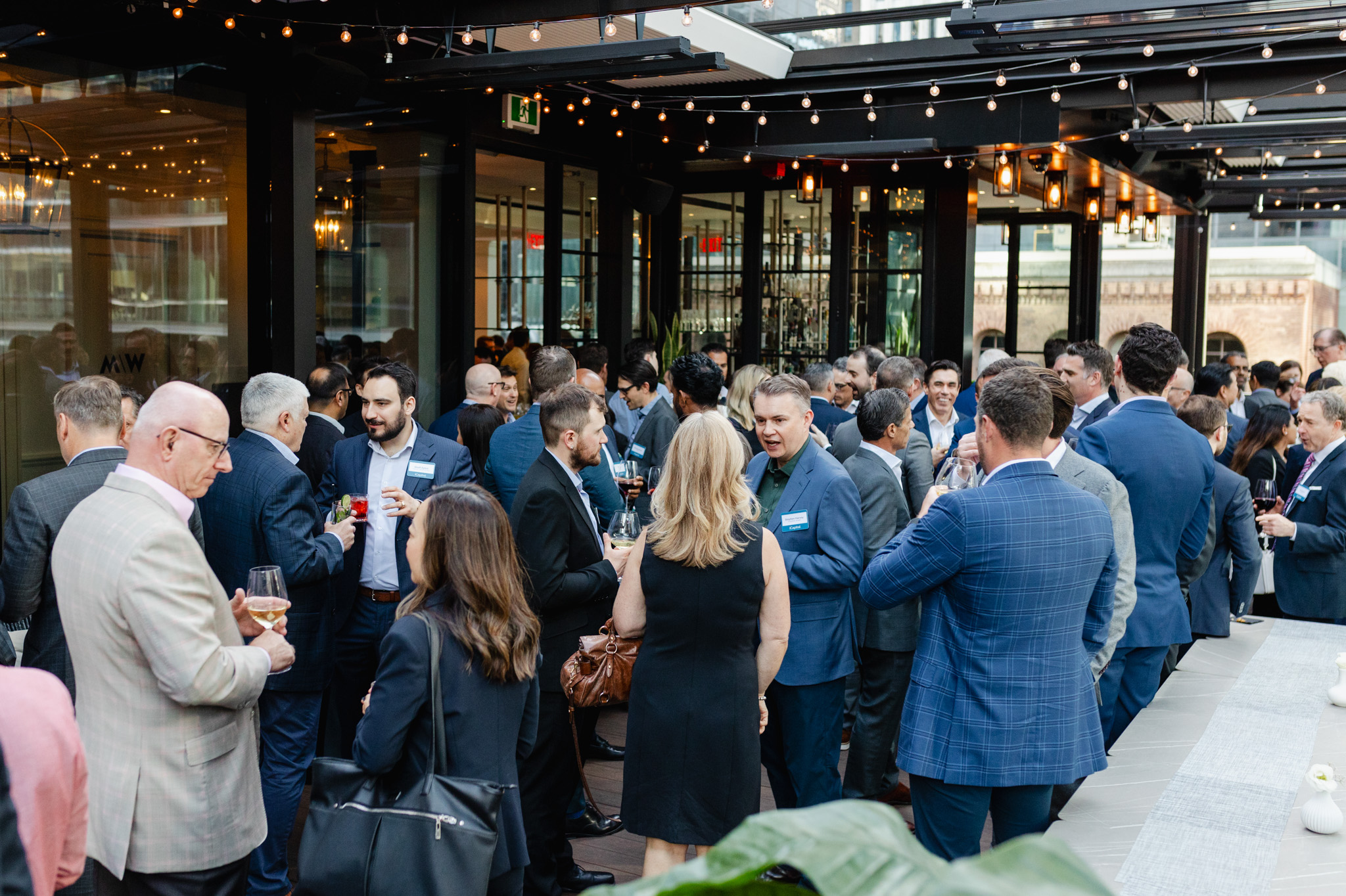 A large group of people in formal attire engage in conversation at an outdoor networking event, with string lights overhead and city buildings visible in the background—a perfect scene for corporate photography.