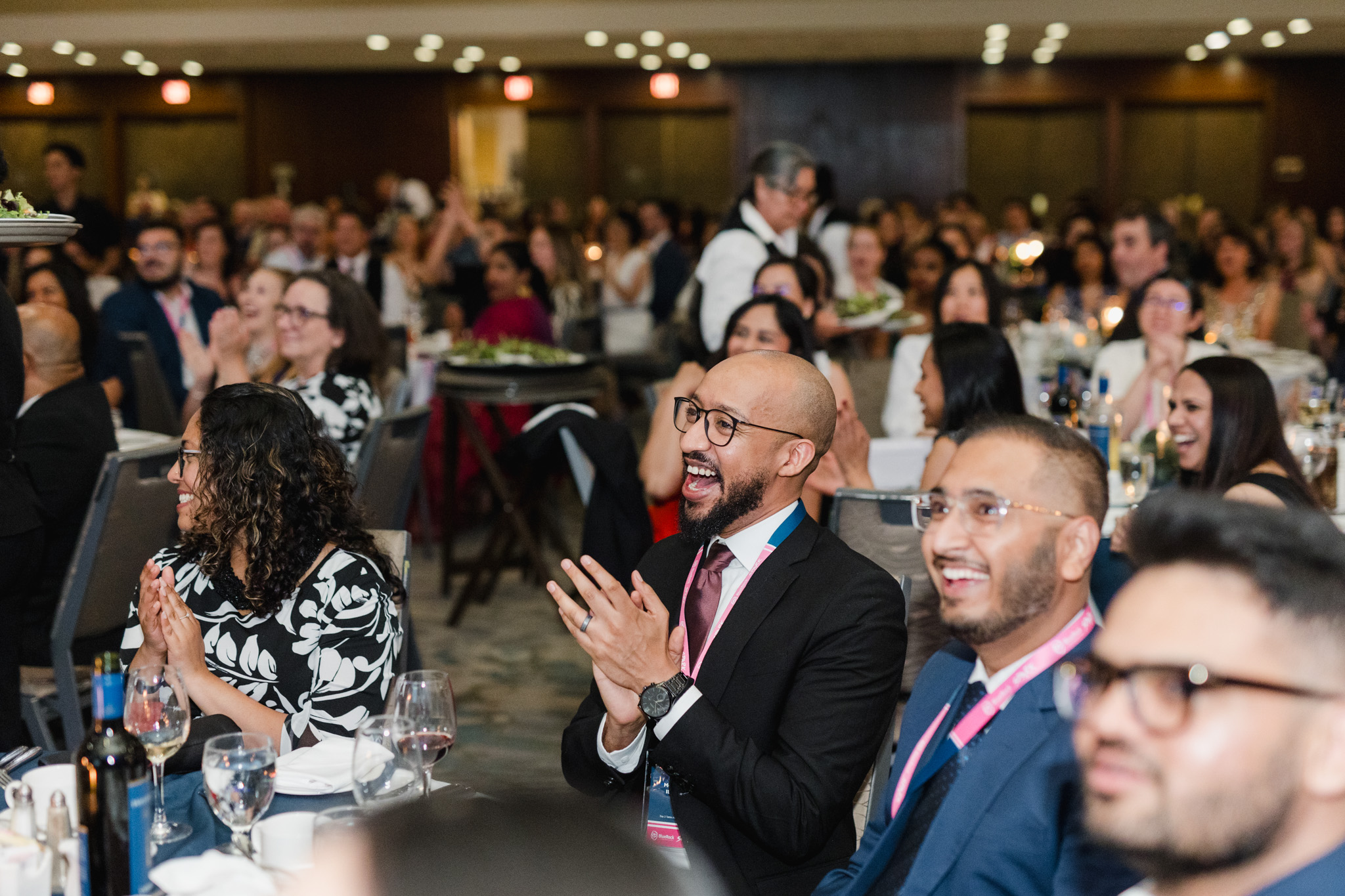 A group of people, seated in a banquet hall, smile and clap during the event. Some have glasses of wine on the table in front of them. The background, captured perfectly in corporate photography style, is filled with more attendees and waitstaff.