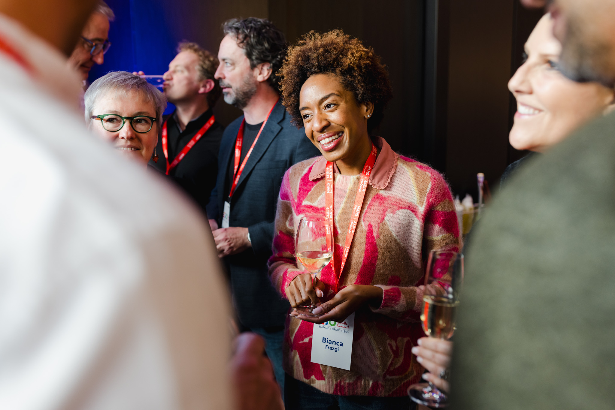 A group of people at a social event. A woman in a pink patterned sweater with a name tag reading "Bianca" is smiling and holding a drink, captured perfectly in this corporate photography session. Others are engaged in conversations around her.