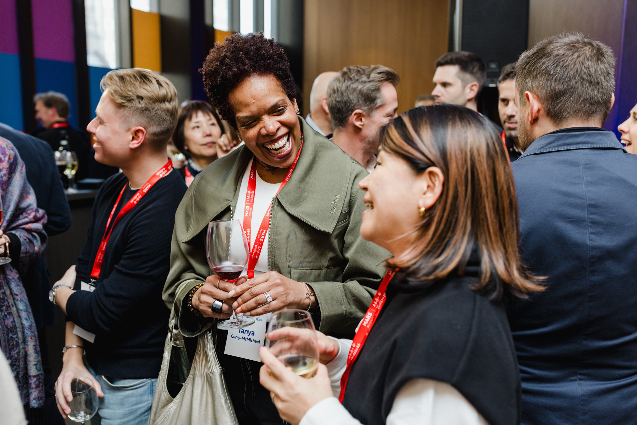 A group of people, wearing ID badges and holding drinks, engage in lively conversation at a networking event, perfect for corporate photography.