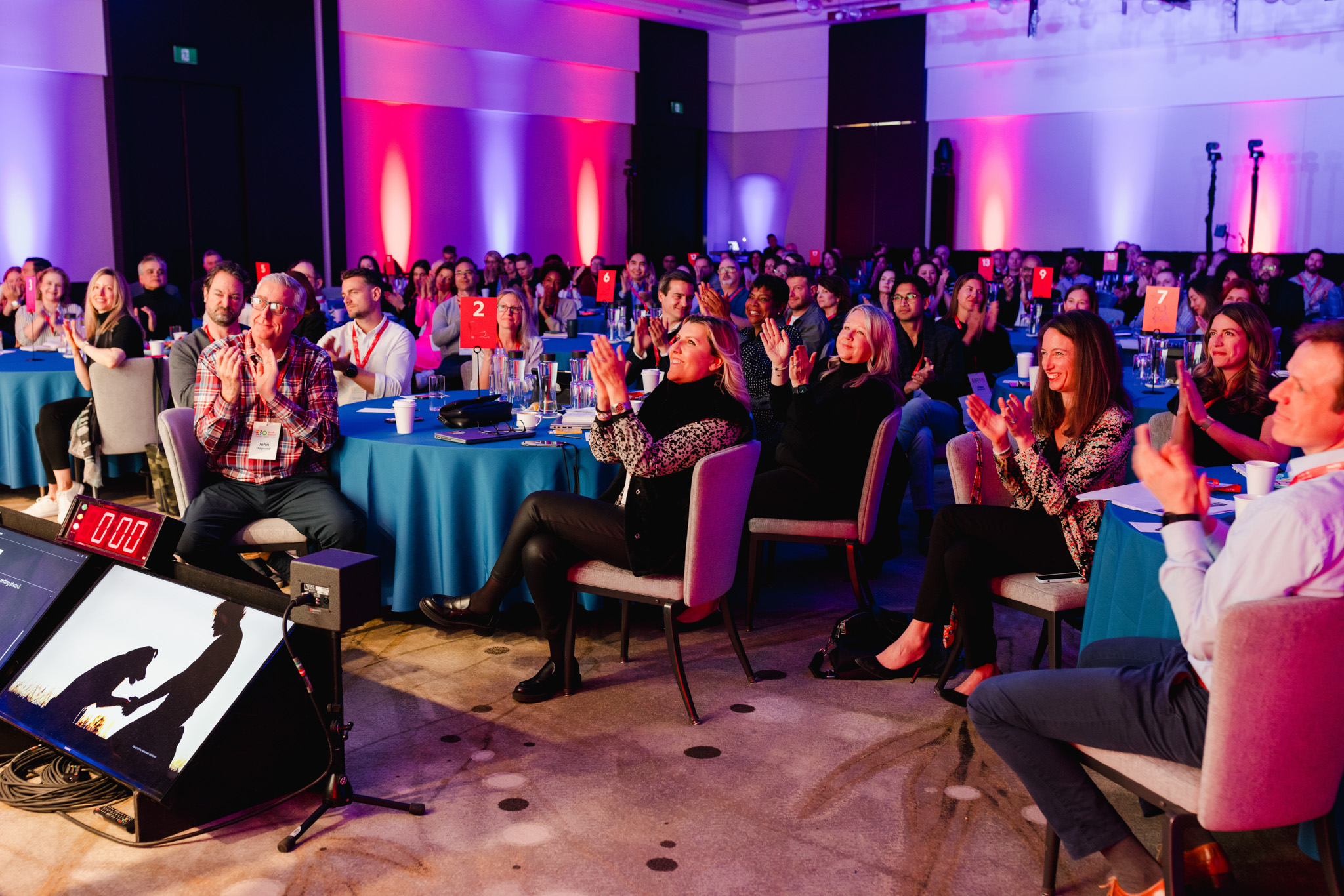 A large audience sits at round tables in a conference room, clapping and facing a presentation screen. The room, captured through corporate photography, is lit with colorful lights in blue and red tones.