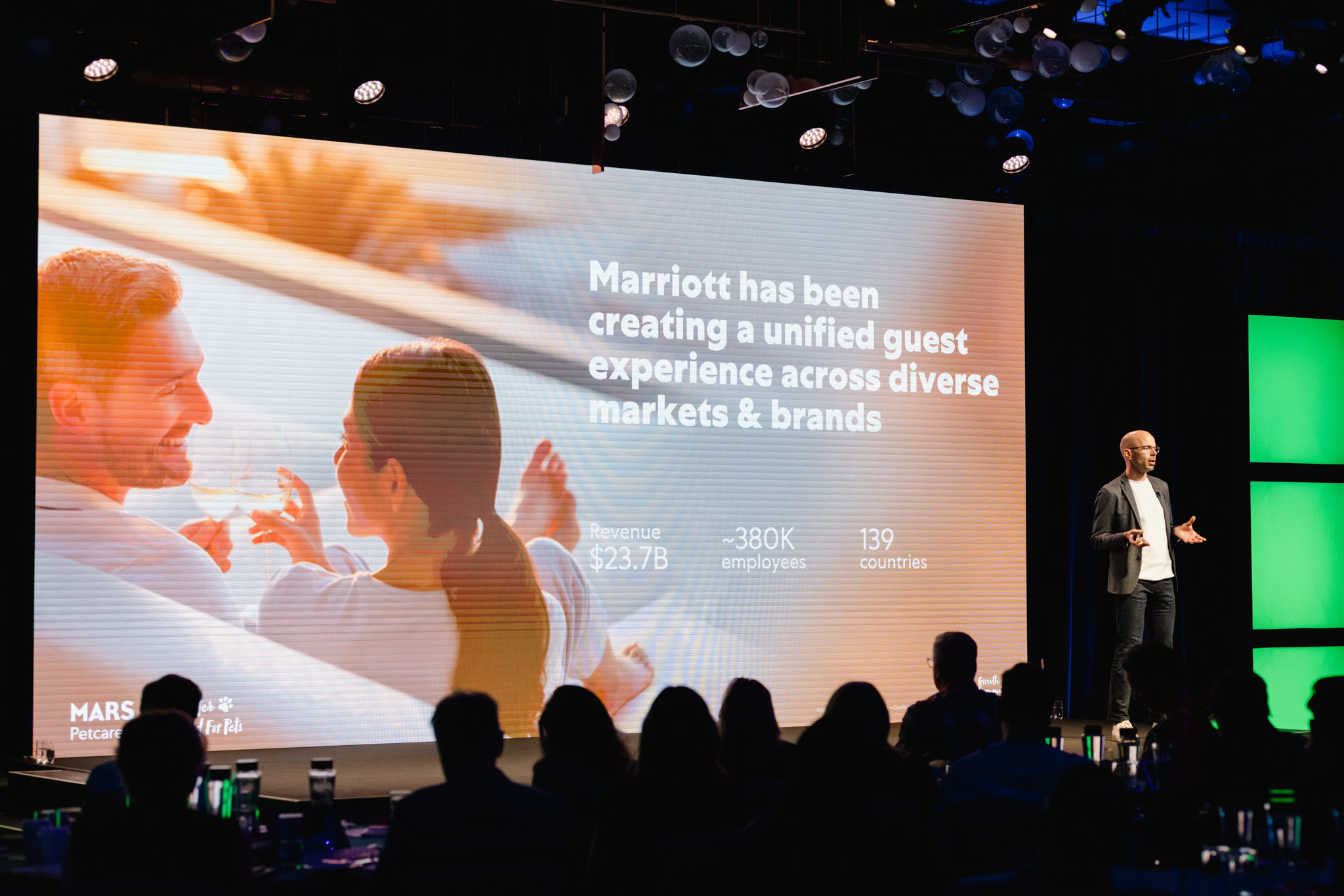A speaker stands on stage next to a large screen with text about Marriott, captured in a moment of corporate photography. The audience is silhouetted in the foreground, while the presentation slide displays revenue, employee count, and operating countries.